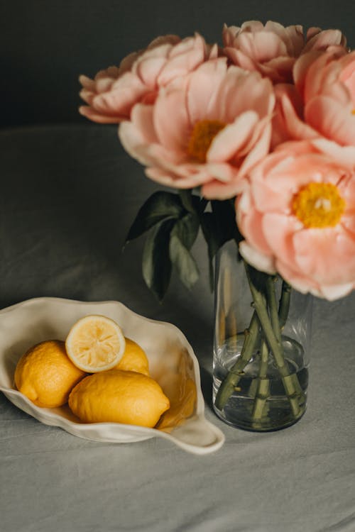Heap of yellow sour lemons placed near glass vase with blooming peony flowers with green stalks on table in light room