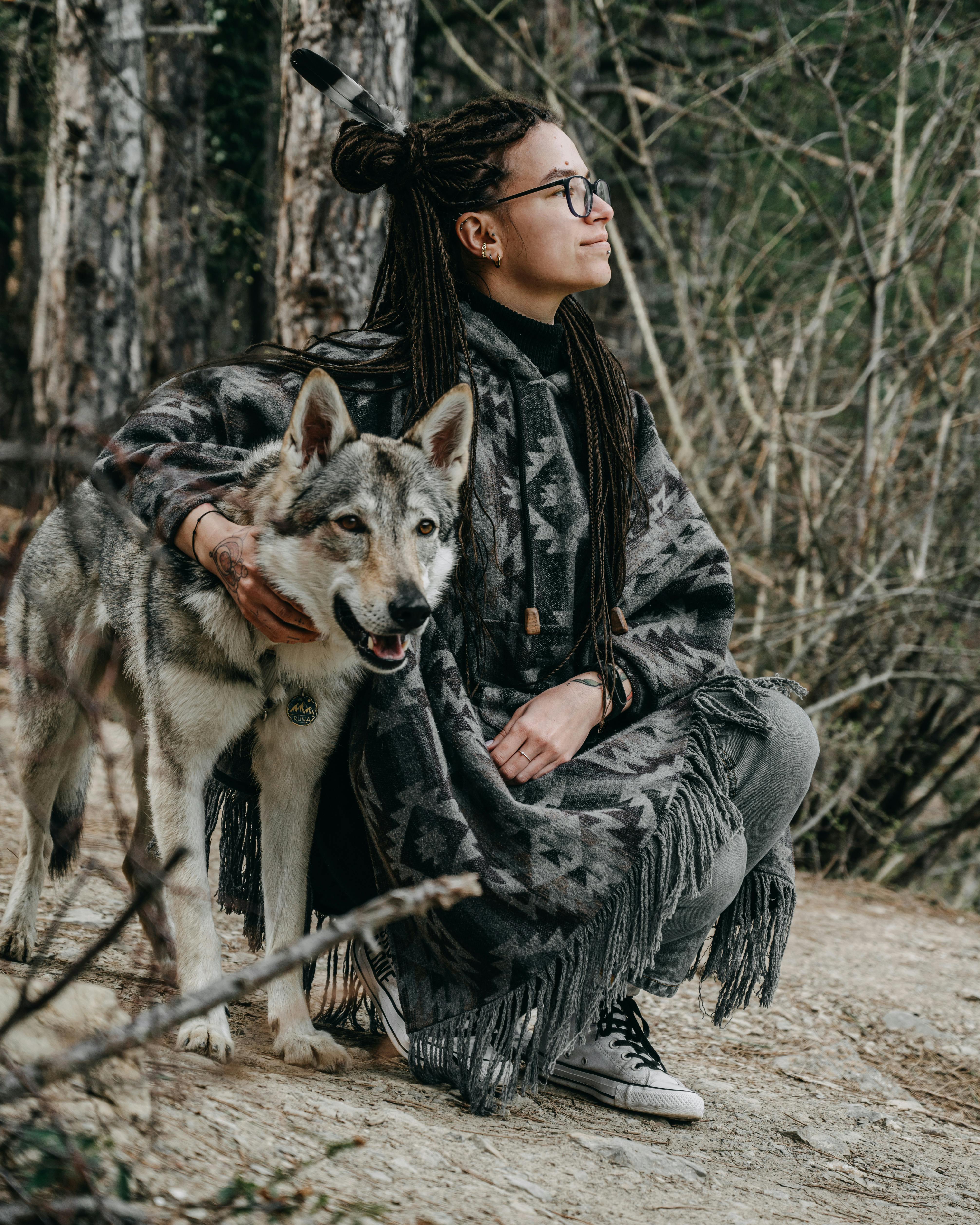 Side view of calm young woman with dreadlocks in stylish poncho squatting down and embracing obedient dog in forest in daytime