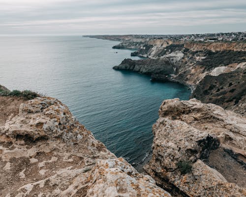 Rough terrain of rocky cliffs washed by rippling water of vast ocean under gray cloudy sky