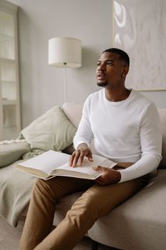 Blind African American man reading braille book on a couch indoors, symbolizing accessibility and education.