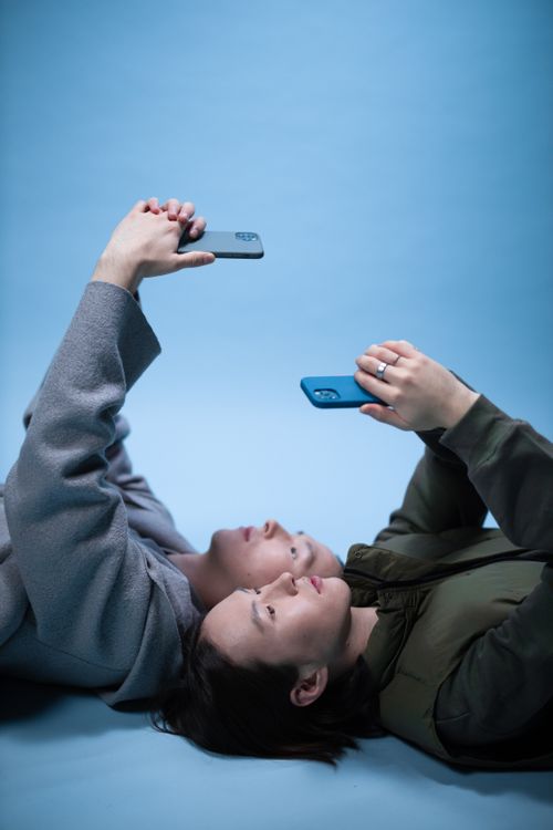 Free Two adults lying down, using smartphones against a bright blue backdrop. Stock Photo