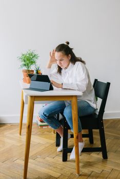 Teenage girl sitting at home desk, learning online with a tablet in a relaxed setting.