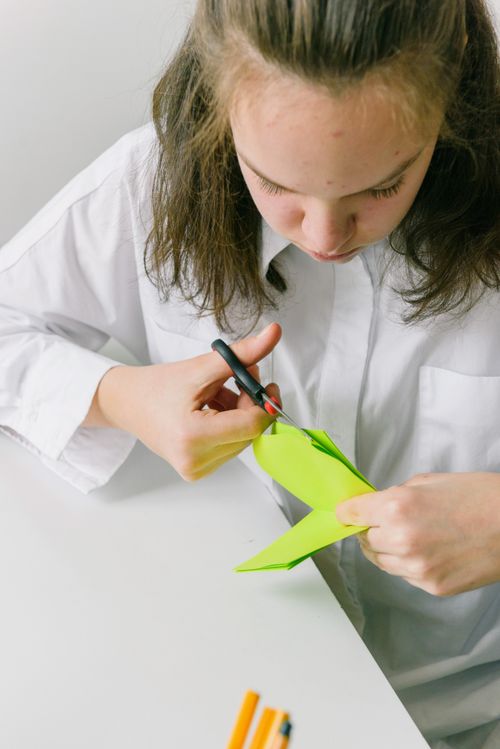Free Teen girl focused on papercraft project, cutting green paper with scissors at a white desk. Stock Photo