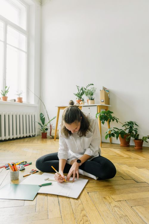 Free A woman is sitting on a wooden floor, sketching with colorful pencils in a bright, plant-filled room. Stock Photo