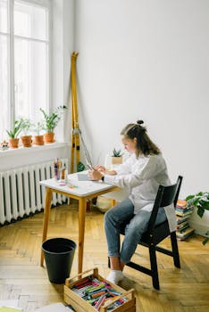 Adult woman focused on a digital project at home, sitting by a window with plants.