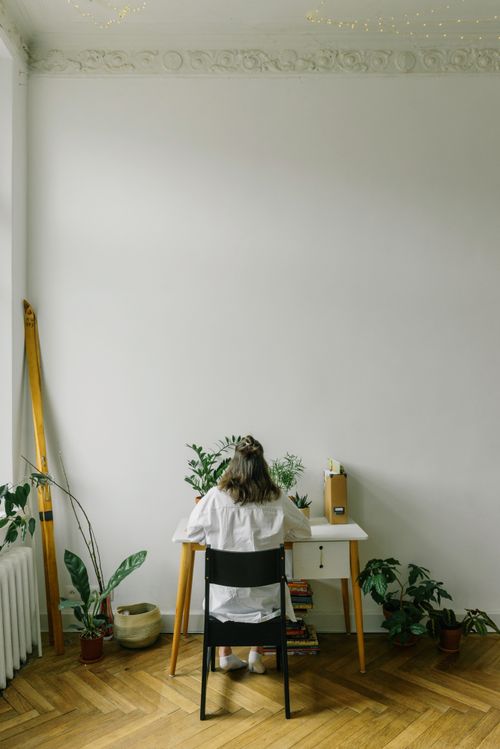 Free A woman sits at a tidy desk surrounded by indoor plants in a minimalist room. Stock Photo