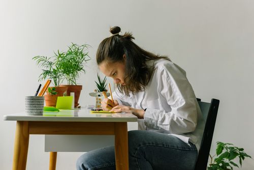 Free A young woman intensely writing at a desk surrounded by potted plants indoors. Stock Photo