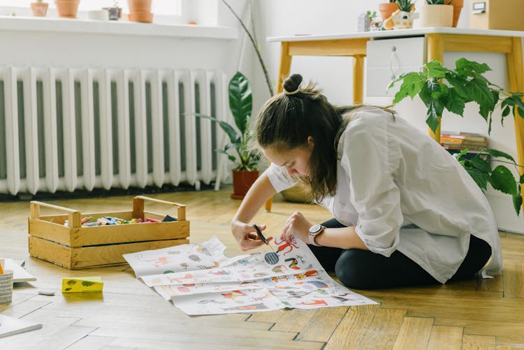 A Woman In White Long Sleeves Sitting On A Wooden Floor While Cutting Paper