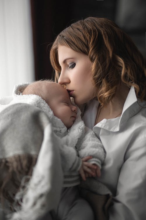 Free Side view of mother kissing warmly dressed newborn baby sleeping in arms at home Stock Photo