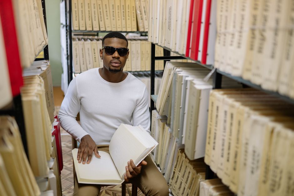 A blind man reading a braille book in a quiet library setting, surrounded by shelves. A blind man reading a braille book in a quiet library setting, surrounded by shelves.
