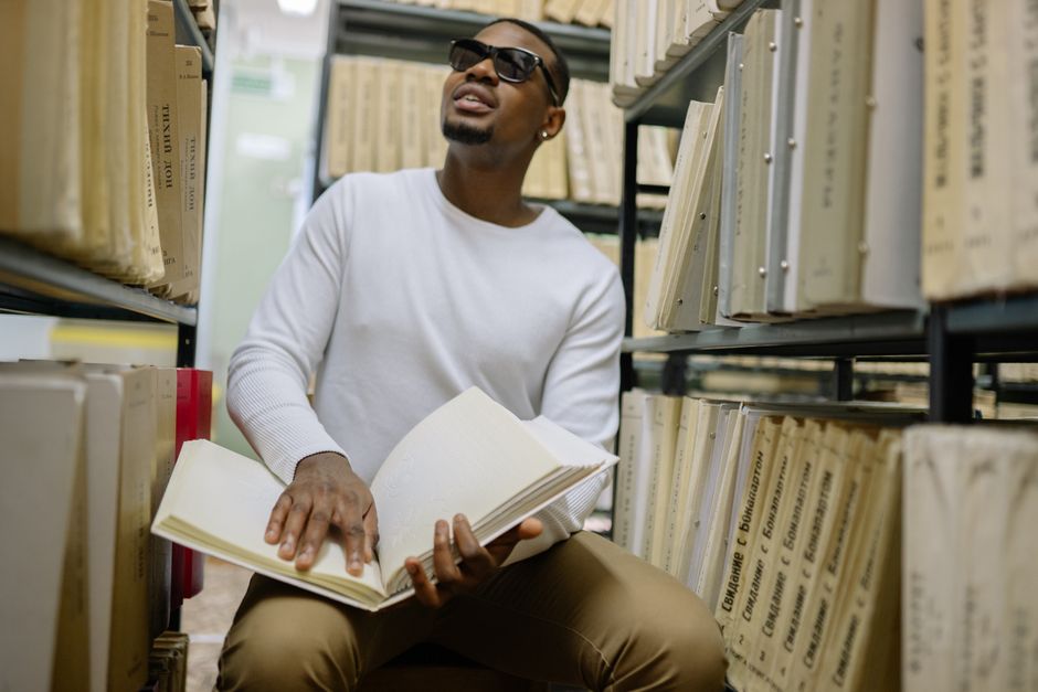 A blind man reads a braille book while sitting in a library surrounded by shelves. A blind man reads a braille book while sitting in a library surrounded by shelves.