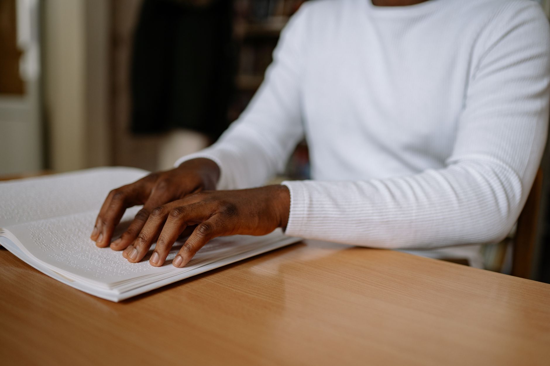 A person using touch to read a Braille book, seated at a desk wearing a white sweater, symbolizing education and literacy.
