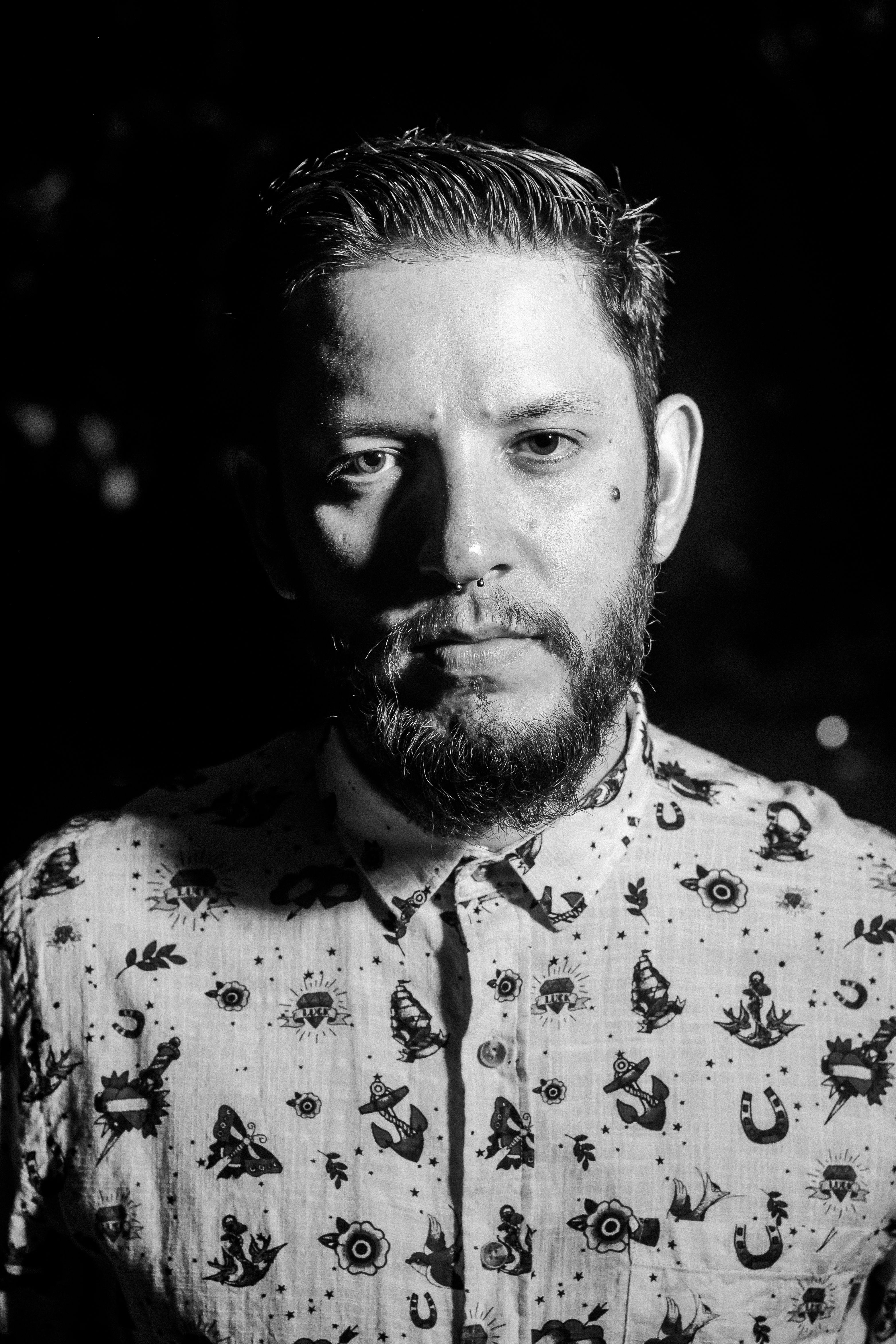Dramatic black and white portrait of a bearded man with unique patterned shirt.