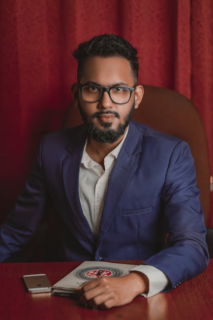 Confident man wearing a blue suit and eyeglasses sits at a desk with a serious expression.