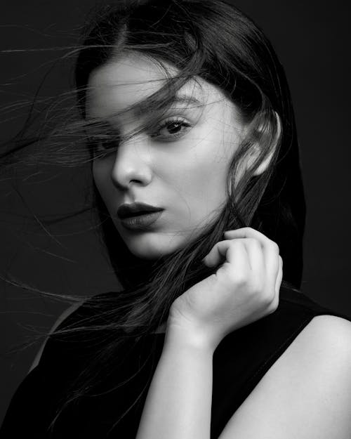 Black and white of young tender female with makeup touching hair while looking at camera