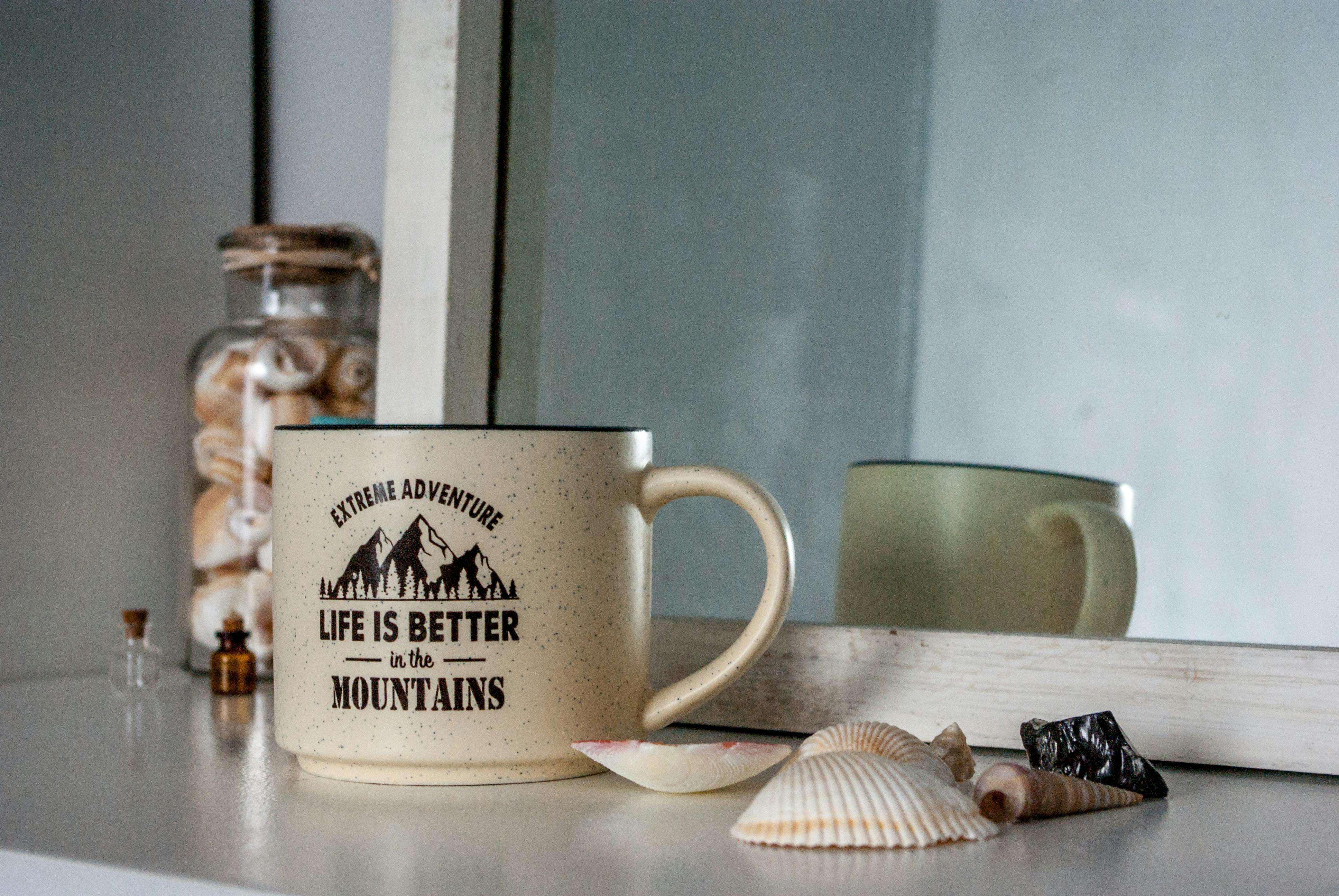 Ceramic mug with mountain quote next to seashells and mirror indoors.