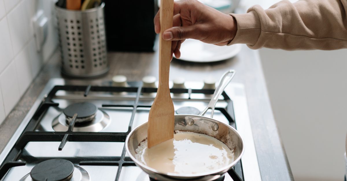Pessoa mexendo molho de creme em uma panela no fogão a gás, usando uma espátula de madeira.