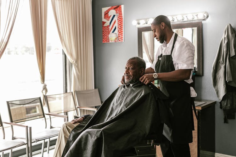 Barber Putting Apron On Mature Black Customer In Barbershop