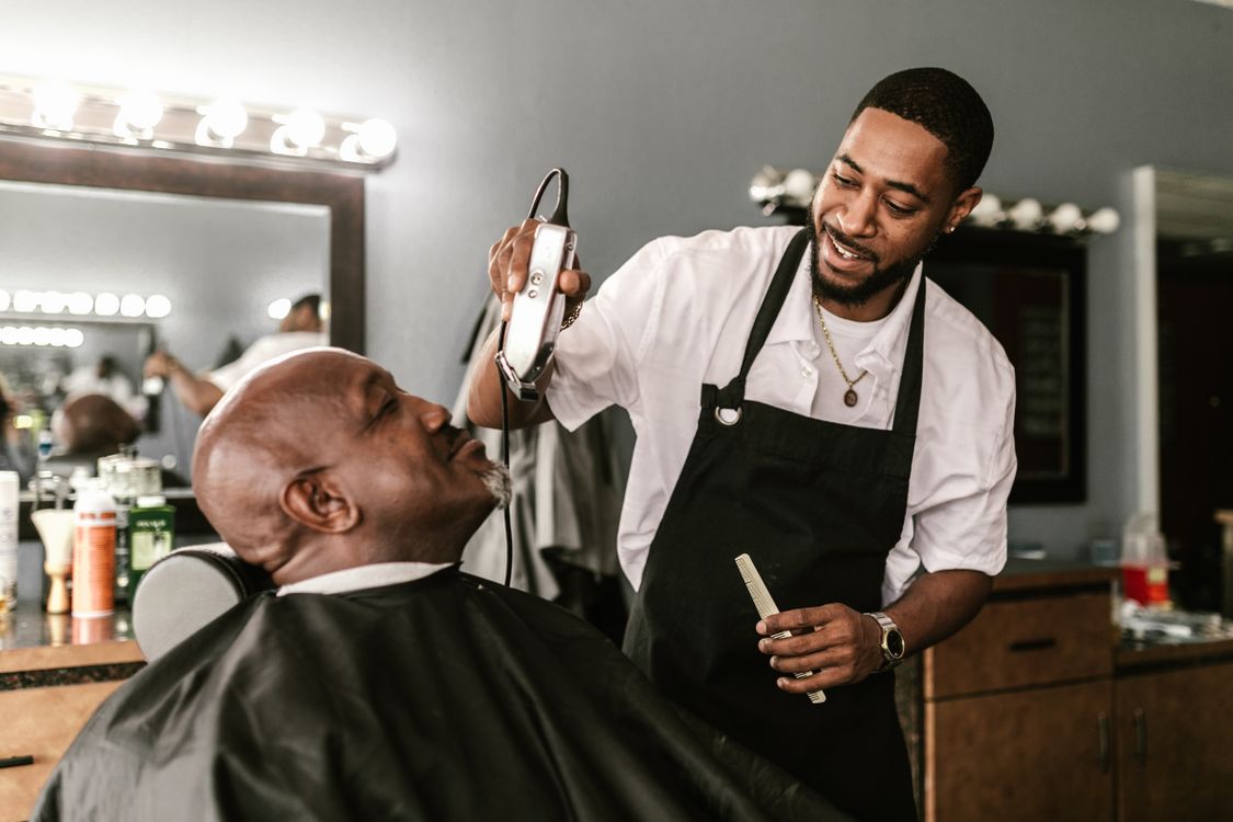Barbeiro usando máquina elétrica para aparar os pelos faciais de um cliente em uma barbearia.