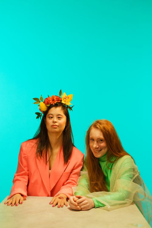 Free Two women sitting at a table, showcasing vibrant fashion with a teal background. Stock Photo