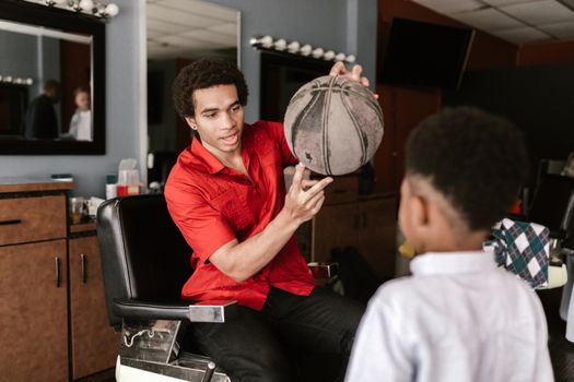 A man sits in a barbershop chair holding a basketball, engaging with a child indoors.