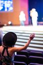 Selective focus of back view of unrecognizable female viewer in hat standing with raised arm in concert