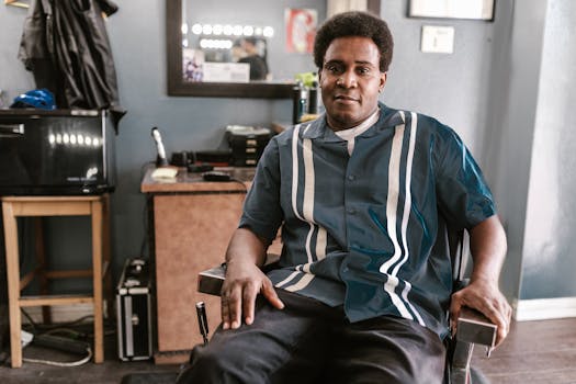 A man in a striped shirt sits smiling in a cozy barber shop, captured in a candid moment.