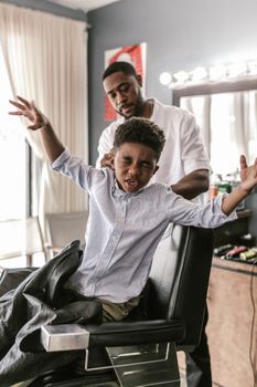 A boy with a playful expression gets a haircut from a barber. Bright indoor scene.