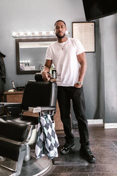 A barber stands confidently next to a chair in a stylish, modern barbershop interior.