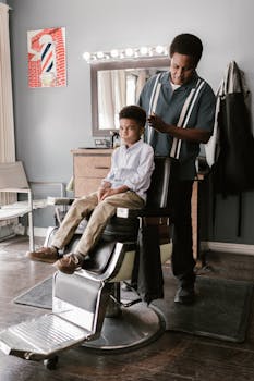 A barber is trimming a young boy's hair in a classic barber shop setting.