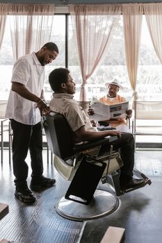 Barber gives a haircut to a client in a contemporary barbershop while another man relaxes with a newspaper.