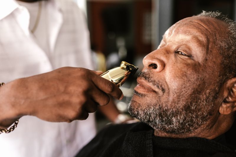 A barber trims an older Black man's beard with clippers in a barbershop.