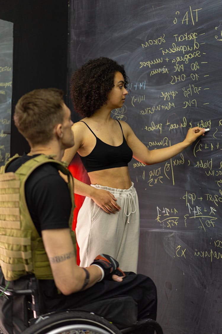 Woman Standing In Black Spaghetti Crop Top Writing On A Blackboard In Front Of A Man Sitting On A Wheelchair