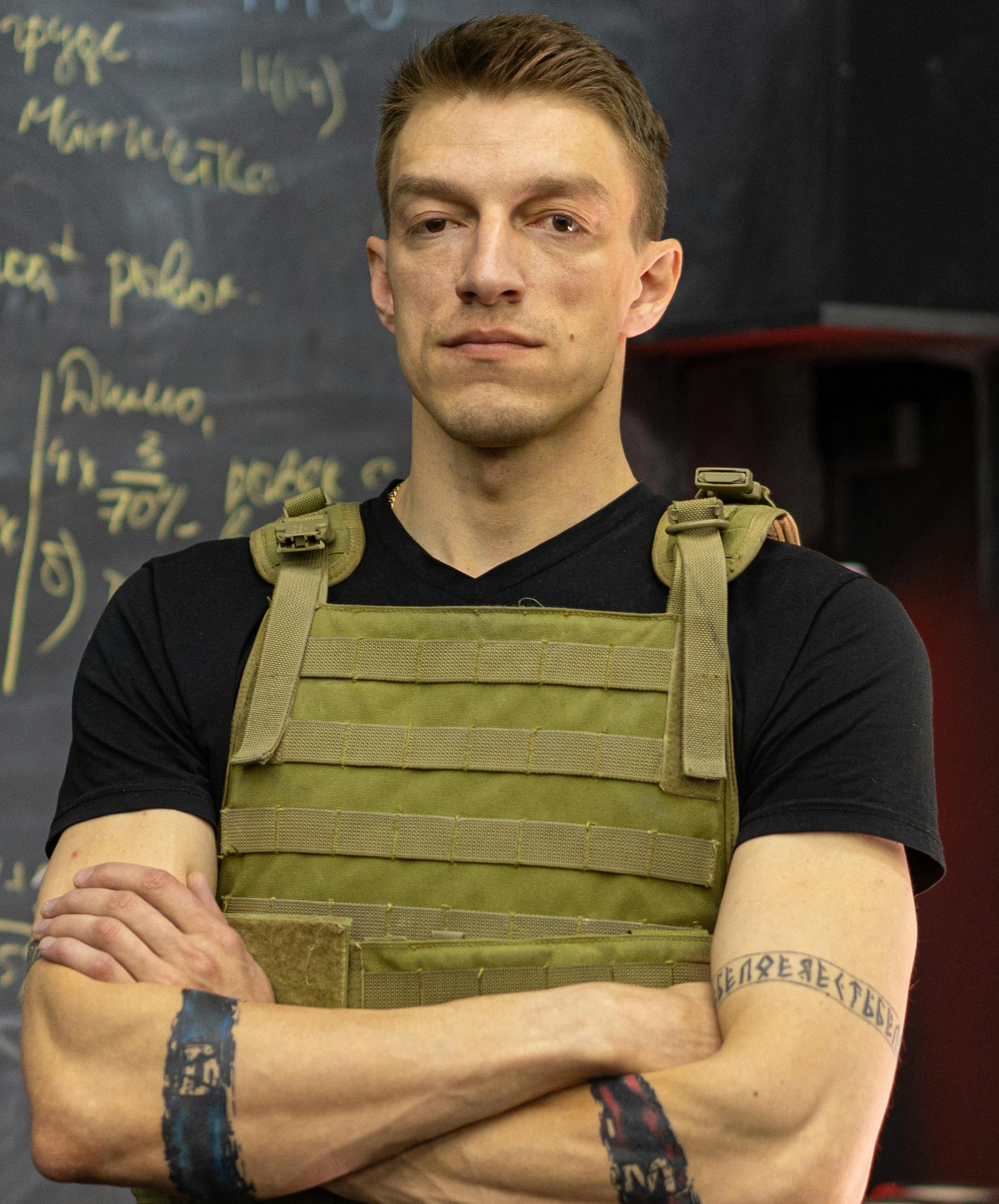 Portrait of a stern man in tactical gear with tattoos, standing against a blackboard background.