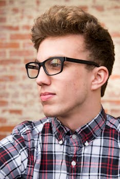 Portrait of a fashionable young man with eyeglasses, wearing a plaid shirt, against a brick wall.