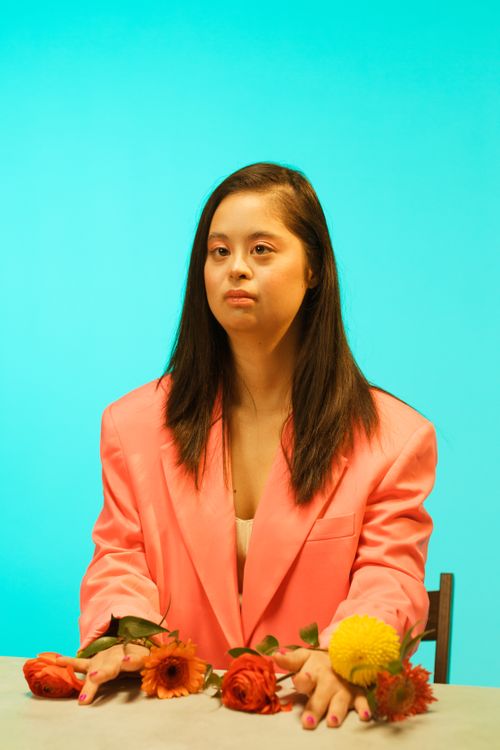 Free A woman in a pink blazer sits against a teal background, surrounded by flowers. Stock Photo