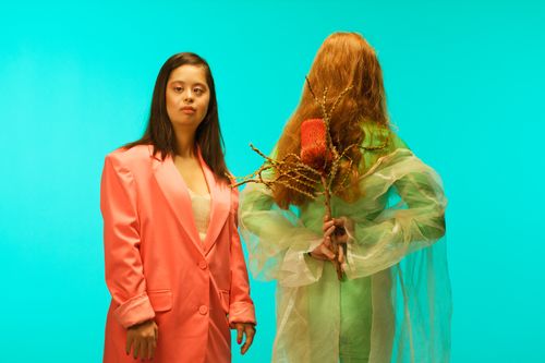 Free Two women posing against teal background, one in a pink blazer, the other holding a dried flower. Stock Photo