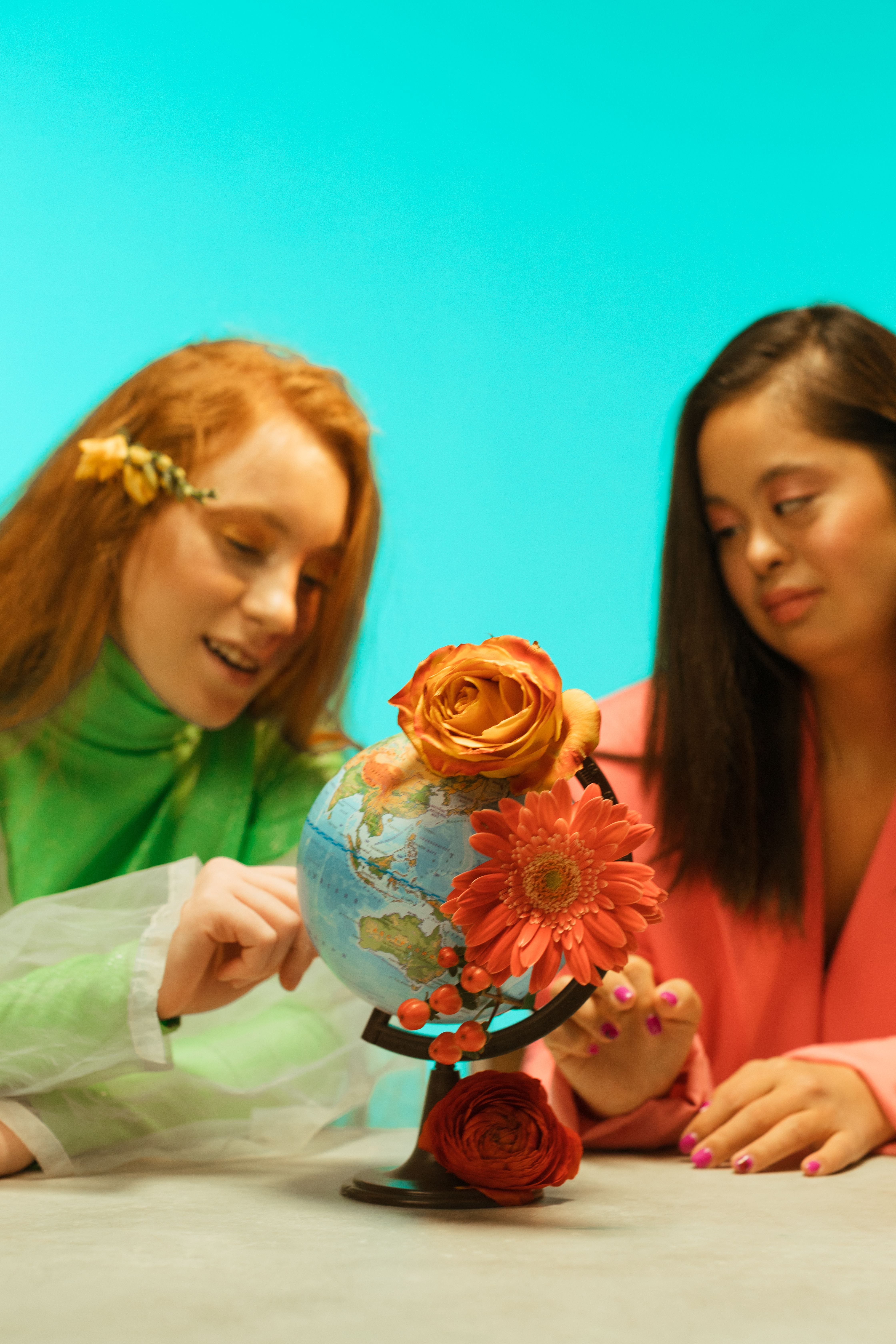 Two women engaged with a decorative globe adorned with flowers on a teal background.