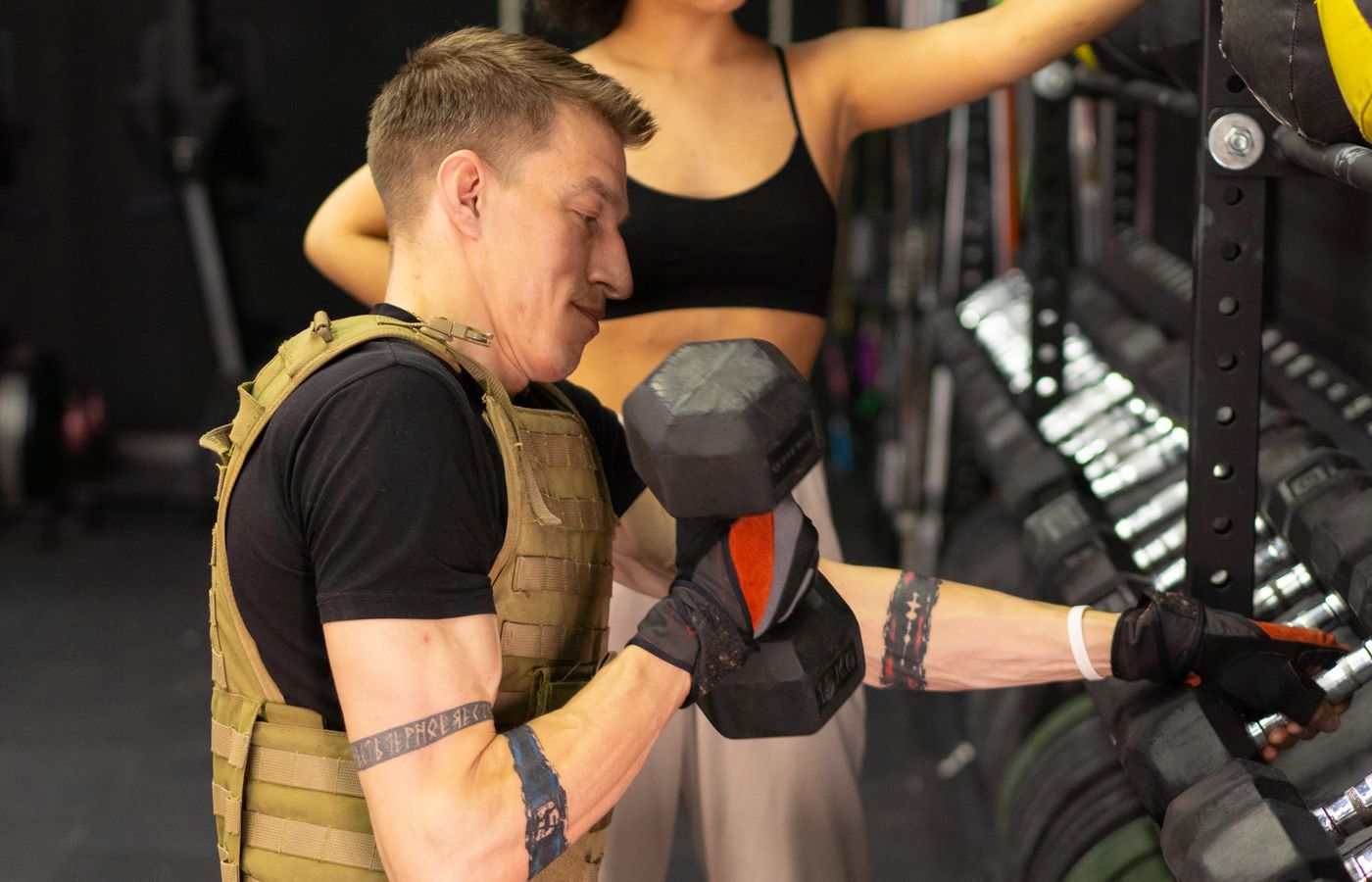 Side view of a man working out with a dumbbell and weighted vest in a gym setting.
