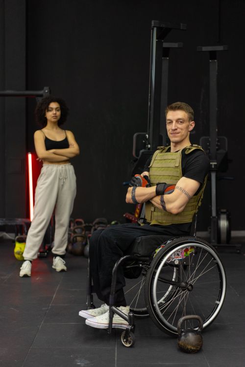 gratis A man in a wheelchair exercising with kettlebells at the gym, assisted by a trainer. Stockfoto