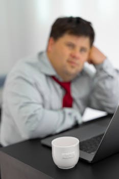 A thoughtful businessman gazes off while seated at a desk with laptop and white mug.