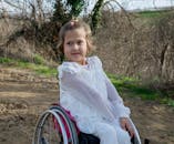 Adorable disabled girl in white clothes looking away while sitting in wheelchair on sandy ground in nature with growing trees