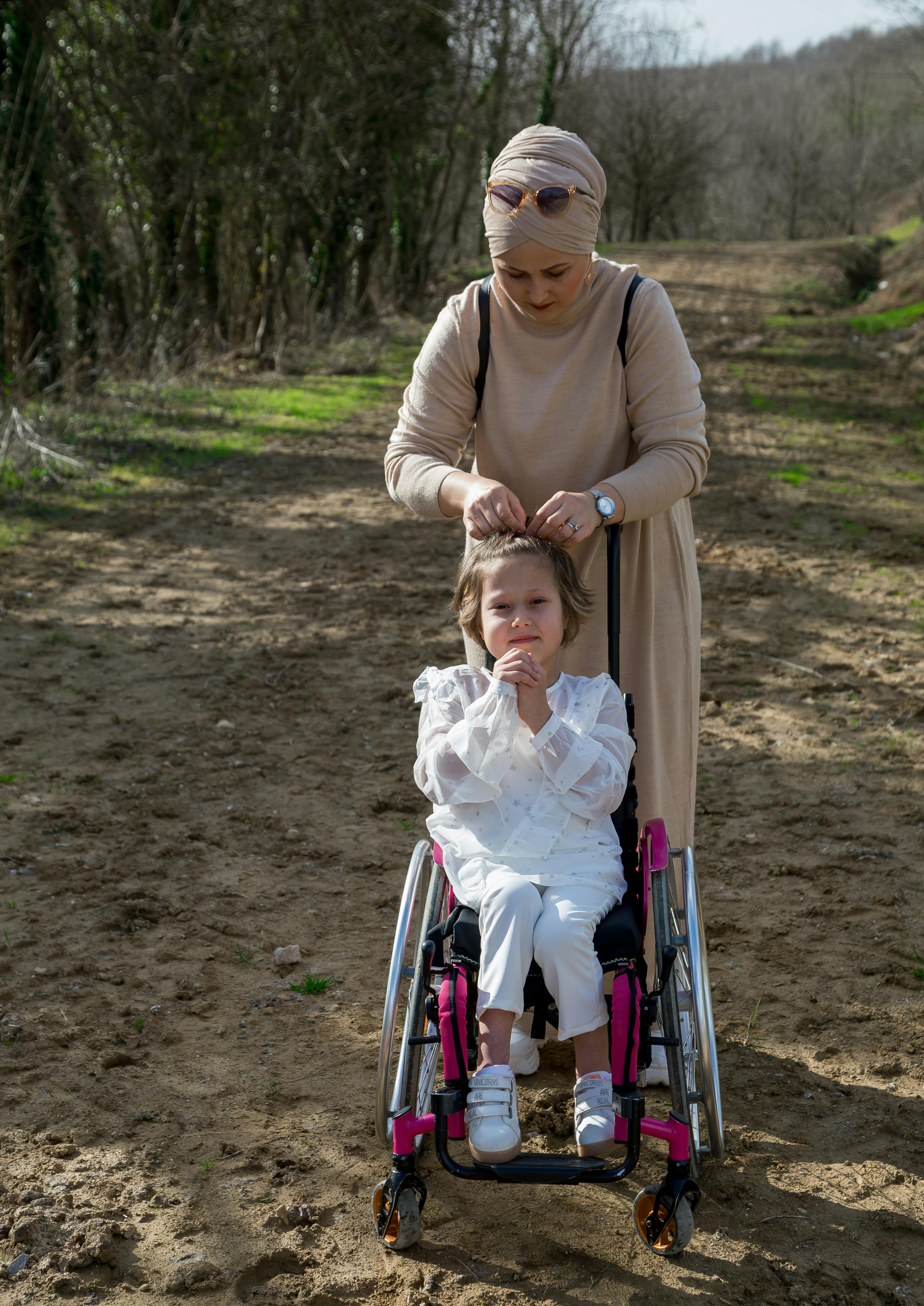 A mother braids her daughter's hair while she's seated in a wheelchair on a forest path.
