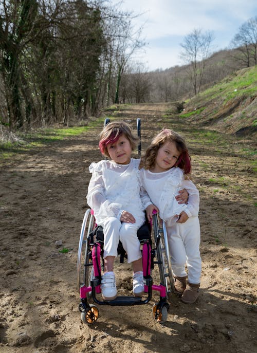 Cute carefree little sisters embracing each other on path surrounded with trees and green grass under cloudy blue sky