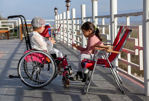 Two young girls enjoying a sunny day on a terrace, one in a wheelchair, sharing a sweet moment.