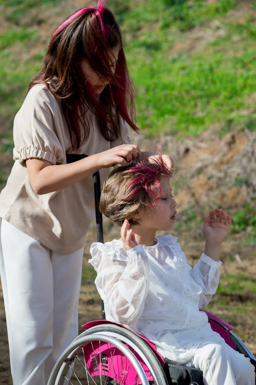 Creative girl touching hair of little sister in wheelchair in park with fresh verdant grass