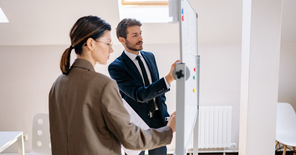 Two colleagues in formal attire discussing strategies on a whiteboard in a modern office space.