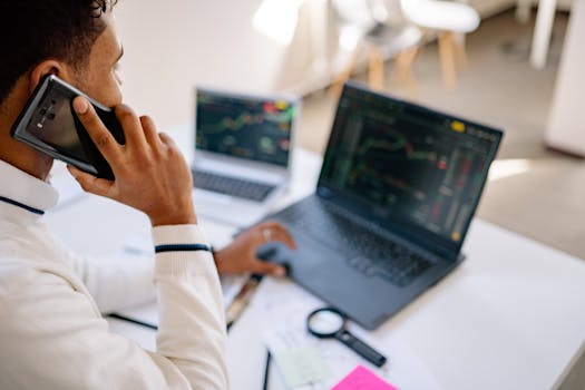 A business professional using a laptop and phone for stock market analysis in a modern workspace.