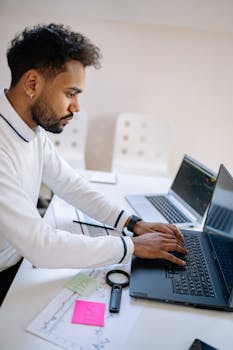 Side view of a man typing on a laptop, analyzing financial data in an indoor office setting.
