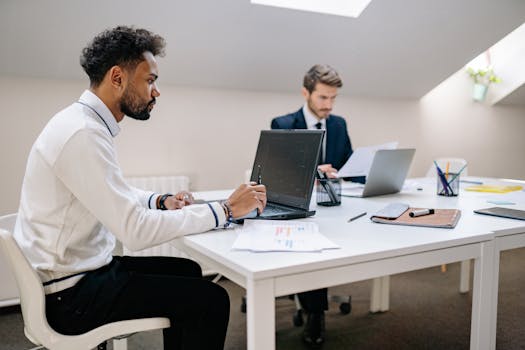 Two men working collaboratively in a modern office setting, focusing on tasks and laptop screens.
